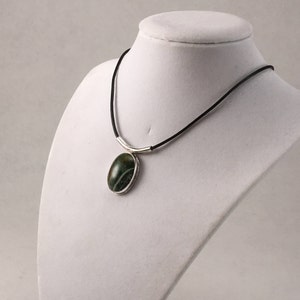 Puede incluir: Un collar de plata con un colgante de &aacute;gata verde en un cord&oacute;n de cuero negro.