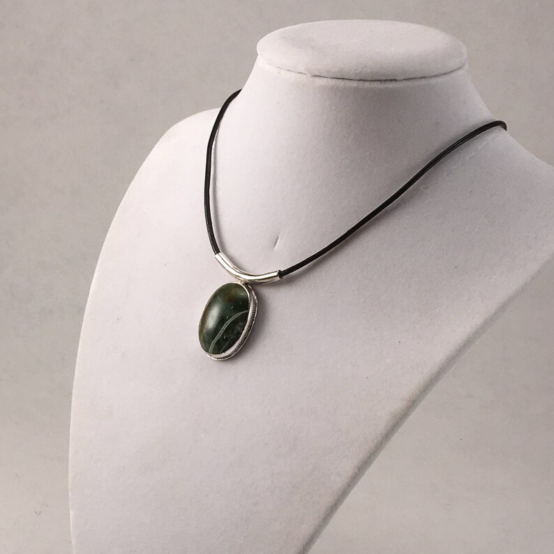 Puede incluir: Un collar de plata con un colgante de &aacute;gata verde en un cord&oacute;n de cuero negro.