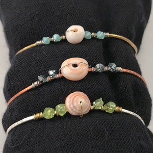 May include: Three handcrafted bracelets with a beach theme. Each bracelet features a natural seashell as the focal point, accented with small beads in shades of blue and green. The bracelets are made with gold, copper, and silver-toned wire.