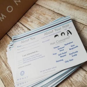 May include: A stack of white cards with blue stripes. The cards have a form for a hair consultation. The form asks questions about hair type, hair concerns, and hair care routine. The form also includes a section for additional comments. The cards are branded with the MONAT logo.