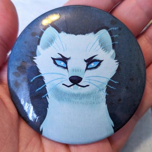 May include: A round button with a white arctic fox with blue eyes on a dark blue background.