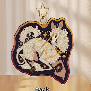 Puede incluir: Un unicornio blanco y esponjoso con un cuerno y una cadena dorados, sobre un fondo azul oscuro con estrellas doradas. El unicornio est&aacute; acostado con la cabeza girada hacia un lado. La imagen est&aacute; en un llavero redondo con borde dorado.