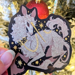 May include: A white unicorn with a gold horn and chain, surrounded by gold stars on a black background. The unicorn is depicted in a stylized, almost cartoonish style.