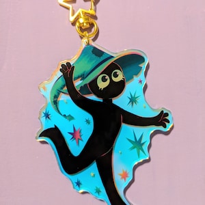 May include: A black silhouette of a person wearing a green witch hat and a blue iridescent background with stars. The person is holding their hat and has a surprised expression on their face. The image is a charm or keychain.