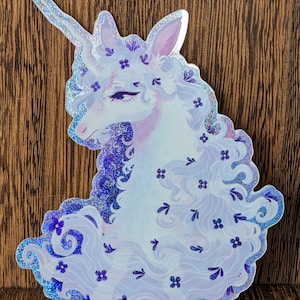 May include: A sticker featuring a majestic unicorn with a flowing mane and a spiraled horn. The unicorn is primarily white with purple accents, adorned with small purple flowers and leaves. The sticker has a shimmering, iridescent border.