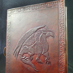 May include: A brown leather journal with a dragon embossed on the front cover. The dragon is detailed with scales and wings. The journal has a decorative border around the edges.