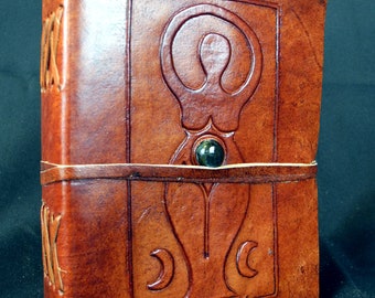 May include: A brown leather journal with a tooled design of a woman with a circle around her head and a crescent moon on each side. The journal has a green stone in the center of the design.