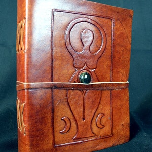 May include: A brown leather journal with a tooled design of a woman with a circle around her head and a crescent moon on each side. The journal has a green stone in the center of the design.