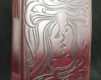 Puede incluir: Un diario de cuero marrón oscuro con un diseño en relieve de dos caras de mujeres con cabello suelto. El diario tiene una encuadernación cosida y un diseño decorativo en la portada.