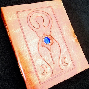 May include: A brown leather journal with a carved design of a woman with a blue gemstone in her belly. The journal has a distressed look and is perfect for writing down thoughts and ideas.