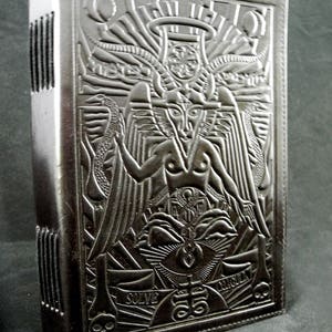 Peut inclure: Journal reli&eacute; en cuir noir avec un motif en relief repr&eacute;sentant une figure ail&eacute;e, un &oelig;il et les mots "Solve" et "Circuit".