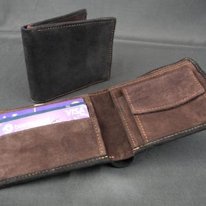 Könnte beinhalten: Ein schwarzes, zweifach gefaltetes Portemonnaie aus Wildleder-ähnlichem Material mit braunem Innenraum und sichtbaren Kartenfächern. Das Portemonnaie ist geöffnet und zeigt das Innere, ein weiteres geschlossenes Portemonnaie ist im Hintergrund.