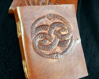 Pode incluir: Dois diários de couro castanho com detalhes dourados. A capa frontal do diário superior apresenta um design em relevo de um nó de cobra. A capa frontal do diário inferior é visível, também com um design de nó de cobra.