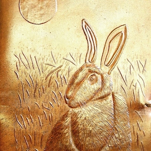 Pode incluir: Uma imagem em relevo dourada de um coelho com orelhas grandes, olhando para cima para a lua cheia. O coelho est&aacute; rodeado por um campo de grama alta.