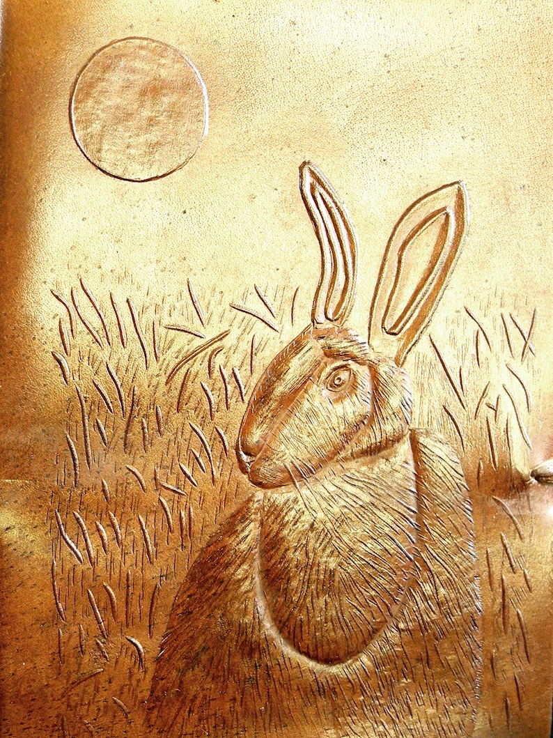 Pode incluir: Uma imagem em relevo dourada de um coelho com orelhas grandes, olhando para cima para a lua cheia. O coelho est&aacute; rodeado por um campo de grama alta.