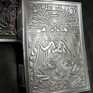Peut inclure: Un journal reli&eacute; en cuir noir avec un motif en relief complexe repr&eacute;sentant une figure ail&eacute;e avec une couronne et un grand &oelig;il. Le texte "SOLVE" et "COGITA" est visible sur la couverture.