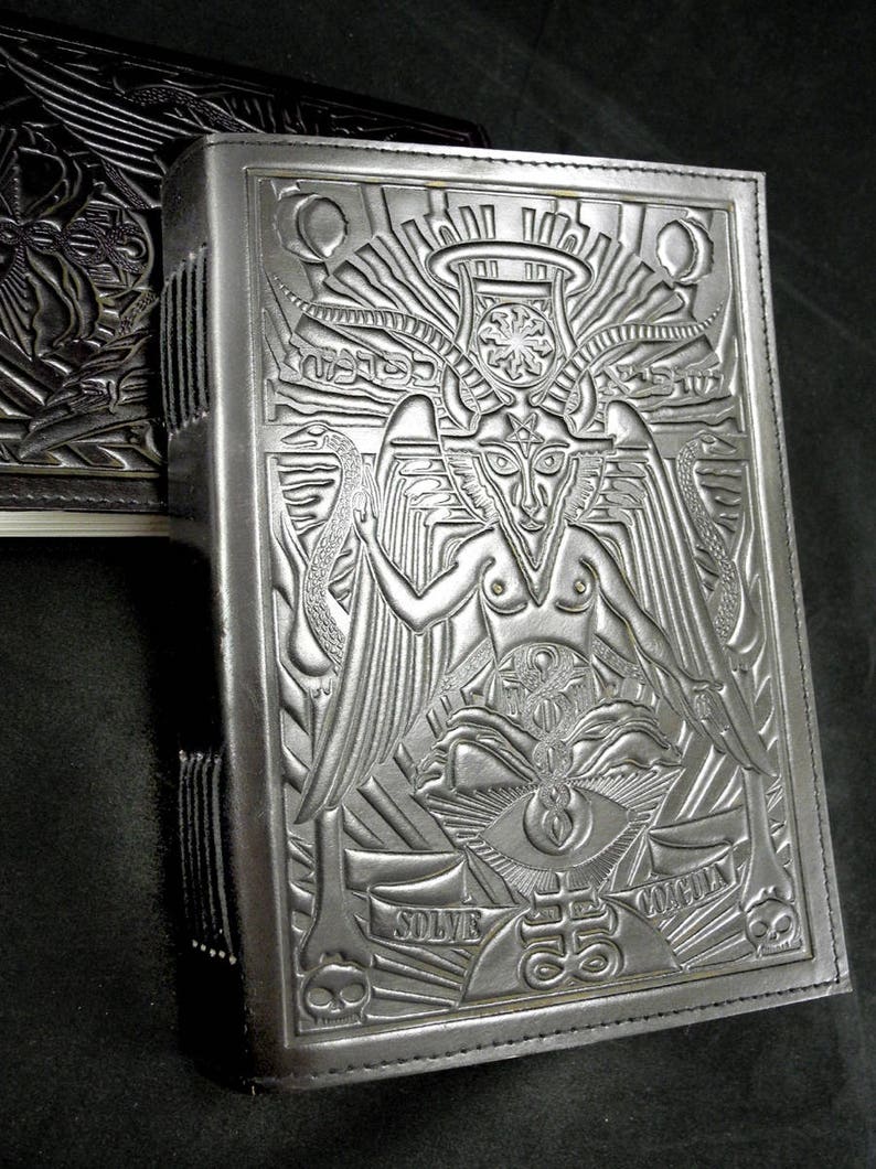 Peut inclure: Un journal reli&eacute; en cuir noir avec un motif en relief complexe repr&eacute;sentant une figure ail&eacute;e avec une couronne et un grand &oelig;il. Le texte "SOLVE" et "COGITA" est visible sur la couverture.