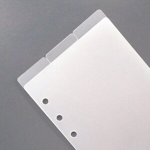 May include: A rectangular, translucent plastic planner divider featuring two top tabs and five circular punched holes along the left side. The divider is made of a smooth, frosted material and is designed to fit into a standard ring-bound planner system.