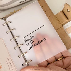 May include: A translucent, frosted plastic planner dashboard featuring the black printed text "Begin with intention." The dashboard is punched with six holes and secured within a tan, textured leather-style ring binder planner.