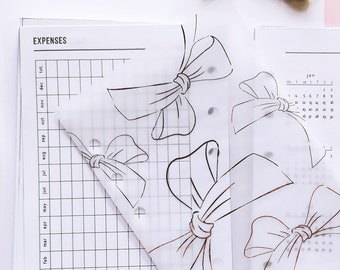 May include: A planner insert set featuring a translucent vellum sheet with copper foil bow illustrations layered over white paper pages. The left page is labelled EXPENSES with a grid, and the right page is labelled PERIOD tracker with calendars.