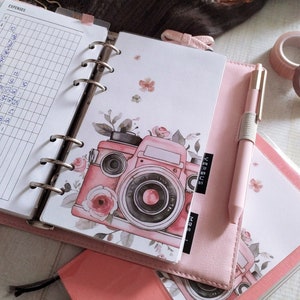 Può includere: Un'agenda a sei anelli rosa con un'illustrazione floreale di una fotocamera sulla copertina. L'agenda include una griglia per le spese, una penna rosa abbinata con dettagli dorati e una linguetta nera con l'etichetta VERSUS.
