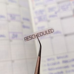 Puede incluir: Una pegatina transparente con la palabra "RESCHEDULED" impresa en negro sobre un fondo blanco. La pegatina está unida a una pequeña herramienta de metal negro.