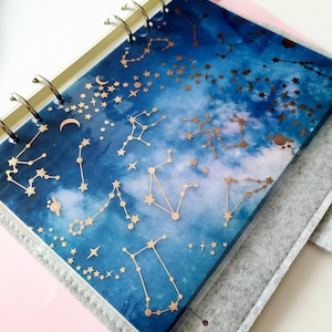 Painel de planejamento em papel vegetal com estampa de céu noturno e constelações, estrelas e planetas em folha de ouro rosa.
