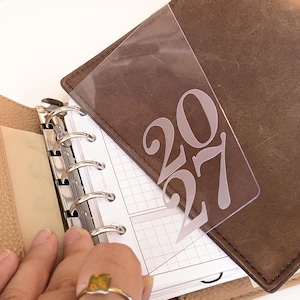 Könnte beinhalten: Eine transparente Kunststoff-Einlage für einen Ringplaner mit der Jahreszahl 2027 in einer großen, stilisierten weißen Schrift. Die Einlage befindet sich in einem braunen Lederplaner mit einem silbernen Sechs-Ring-Bindemechanismus.