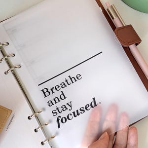 May include: A brown leather ring-bound planner featuring a translucent vellum divider page with the black printed text "Breathe and stay focused." A light pink pen with a metallic silver clip is secured in the integrated pen loop on the side.