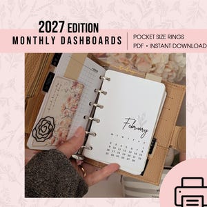 May include: A tan leather pocket-size ring planner open to a February 2027 monthly calendar page. The page features a floral illustration, the text February, and a grid calendar. A decorative card with floral art and a black wire rose paperclip are visible.