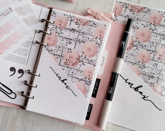 May include: A pink ring-bound planner featuring floral patterned pages with a wood grain background. The planner includes white pockets labeled with the word inbox in black script, a black paperclip, a black pen, and a sheet of pink flag stickers.