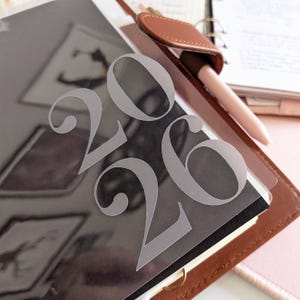 May include: A clear plastic overlay with the numbers "2026" in a light gray, placed over a planner. The planner is brown leather with a pen and a leather pen holder. The background is a light pink.