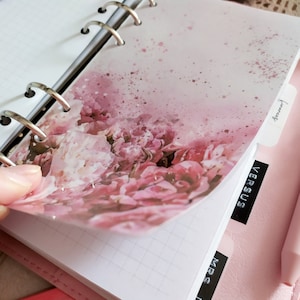 May include: A pink floral patterned planner with a clear plastic cover. The planner has a ring binder and tabs labeled "MRS", "VERSUS", and "Finances".