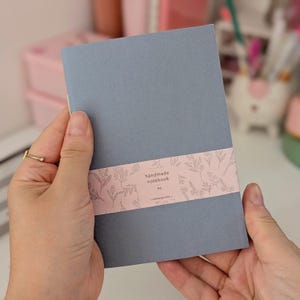 Peut inclure: Un carnet A6 fait à la main avec une couverture grise et une bande florale rose. La bande porte l'inscription "handmade notebook" et "venusprints". Le carnet est tenu à deux mains.