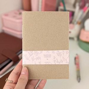 May include: A handmade notebook with a kraft paper cover. A pink band with a floral design wraps around the notebook, displaying the text "handmade notebook" and "A6". The notebook is held by a hand.