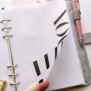 May include: A ring-bound planner with white lined paper and a grey felt cover. A page is partially turned, revealing black printed text on the underside. A pink pen is held in an elastic loop on the cover, and a gold metal binder clip sits nearby.