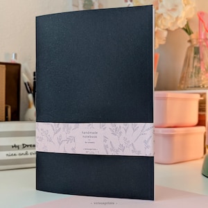 Puede incluir: Un cuaderno negro con una faja de papel rosa claro que presenta un patrón de líneas botánicas en blanco. La faja incluye el texto: handmade notebook, 60 sheets, versusprints. El cuaderno tiene una cubierta negra sólida y forma rectangular.