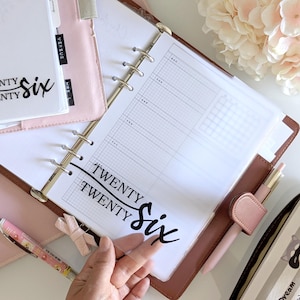 Può includere: Un'agenda con copertina in pelle marrone e anelli dorati, aperta su una pagina a righe. Il testo "Twenty Six" è stampato in nero. Sono visibili anche una penna rosa e un fiocco rosa.