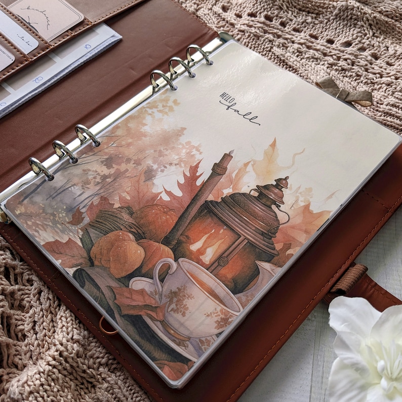 K&ouml;nnte beinhalten: Ein brauner Leder-Ringbuchplaner mit einer dekorativen Einlage mit dem Text Hello Fall. Die Illustration zeigt eine leuchtende Laterne, eine Teetasse und Herbstbl&auml;tter in Orange-, Braun- und Cremet&ouml;nen vor einem neutralen Hintergrund.