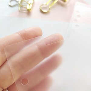 May include: A translucent, frosted plastic divider sheet with a smooth, matte finish. The rectangular sheet features a single circular hole punched near the corner and a small tab extending from the top edge, designed for use in a binder system.