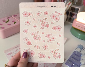 Pode incluir: Uma folha de autocolantes retangular com várias ilustrações de flores de cerejeira cor-de-rosa sobre um fundo de grelha pálido. No centro superior apresenta o texto versusprints com ícones de redes sociais e o site versusprints.com.