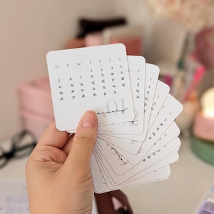 May include: A hand holds a stack of white, square calendar cards with rounded corners. Each card displays a month's calendar in a minimalist style. The January card is visible at the front, with dates in a simple font.