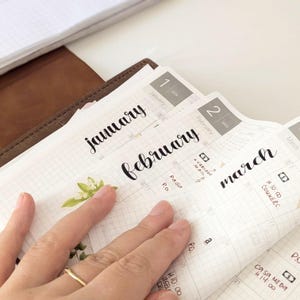 Pode incluir: Uma agenda de 2025 com páginas de papel quadriculado branco mostrando os meses de janeiro, fevereiro e março em escrita cursiva preta. As páginas apresentam notas manuscritas e pequenos adesivos florais, com uma capa de couro marrom visível.