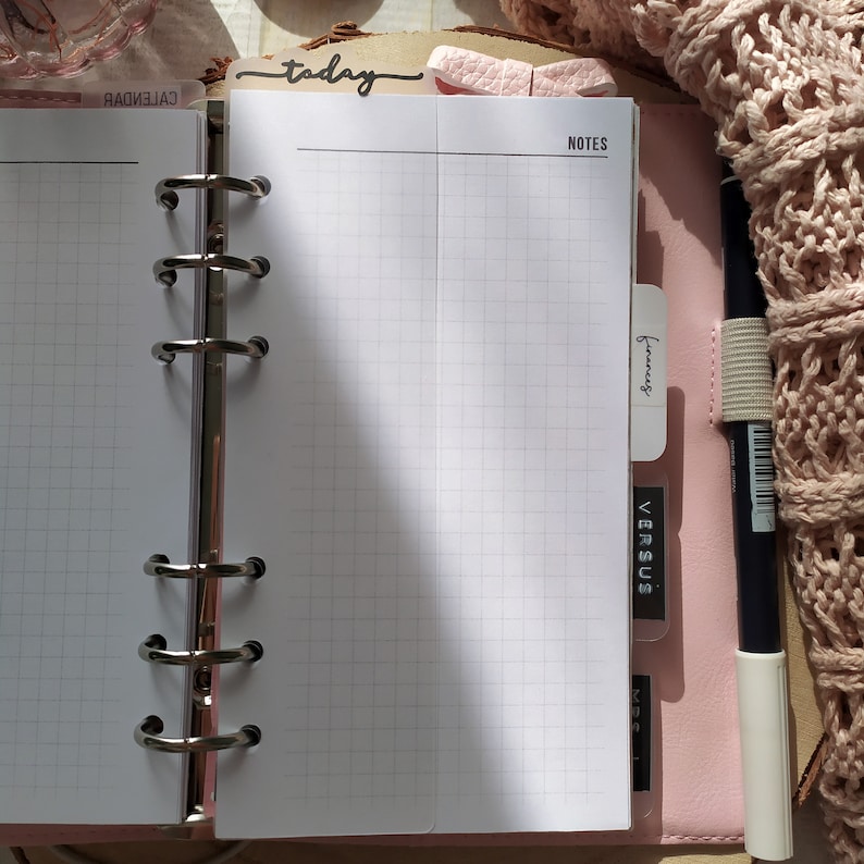 May include: A pink planner with grid paper pages, open to a blank page labelled "NOTES". A black pen with a white tip is secured with a beige elastic band. The word "today" is visible on a tab. The planner is on a wooden surface.
