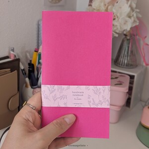 May include: A bright pink rectangular notebook featuring a light pink paper belly band with a white floral pattern. The band includes the printed text handmade notebook, 60 sheets, and versusprints. The notebook has a smooth, solid cover.