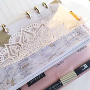 May include: A clear plastic planner bookmark featuring a white intricate mandala cutout design. The bookmark is placed inside a ring-bound planner with visible weekly calendar pages, tabbed dividers labeled Finances and Personal, and a black pen.