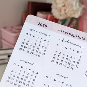 以下が含まれることがあります： 大理石のような背景の2026年カレンダーステッカーのクローズアップ。カレンダーには1月、2月、4月の月が表示され、曜日と日付が黒で印刷されています。「versusprints」というテキストも表示されています。