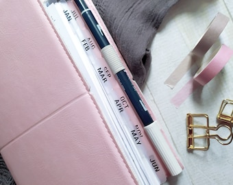 May include: A light pink planner with monthly tab dividers labeled JAN, FEB, MAR, APR, MAY, JUN, JUL, AUG, SEP, OCT, NOV. A dark blue and white dual-tip marker rests on the pages. Two rolls of adhesive tape and two gold metal binder clips are nearby.