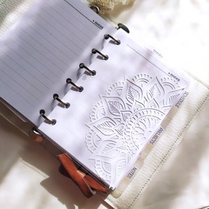 May include: A white ring-bound planner with lined paper, featuring a decorative die-cut mandala pattern on the front page. The planner includes tabbed dividers labeled notes, projects, and schedule, with a brown leather bow accent attached.