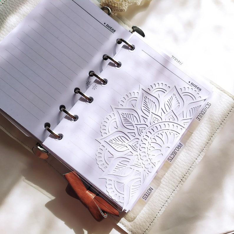 May include: A white ring-bound planner with lined paper, featuring a decorative die-cut mandala pattern on the front page. The planner includes tabbed dividers labeled notes, projects, and schedule, with a brown leather bow accent attached.